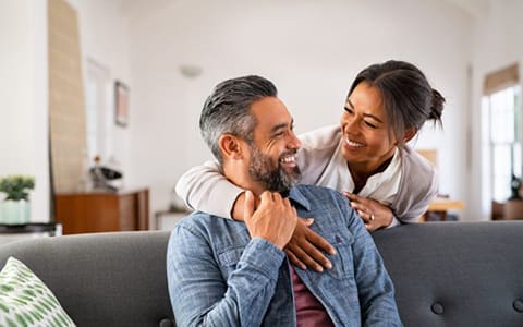 Smiling ethnic woman hugging her husband on the couch from behind in the living room.